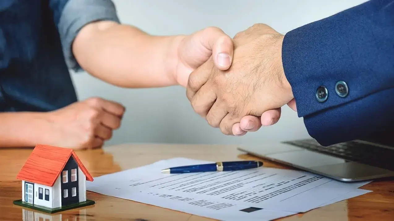 Dois homens apertam as mãos sobre um contrato para fechar um negócio imobiliário, com uma casa em miniatura na mesa.