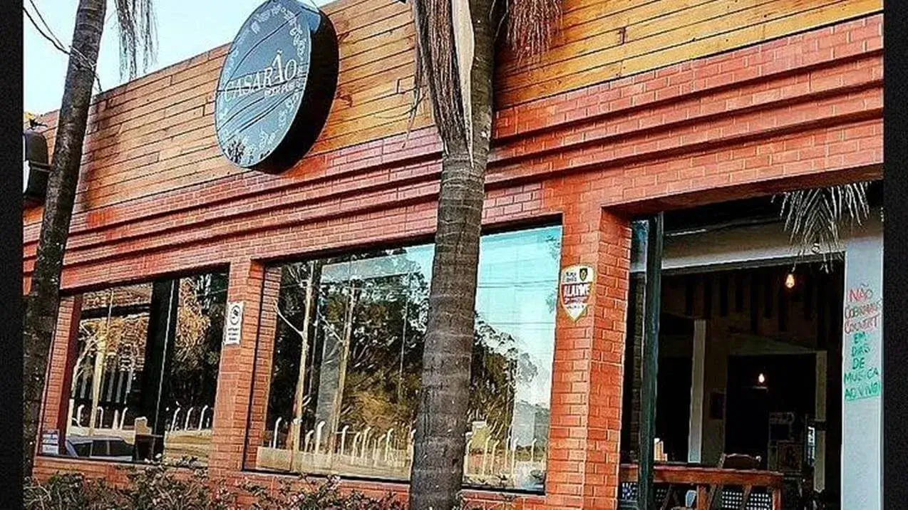 A fachada do Casarão Beer Pub, localizado no bairro de Santa Mônica, em Uberlândia, apresenta uma charmosa combinação de tijolos alaranjados e revestimento de madeira clara na parte superior, criando uma estética convidativa. No lado esquerdo da fachada, destaca-se uma placa circular preta, onde se lê "CASARÃO" em letras brancas, indicando o nome do estabelecimento. Amplas janelas retangulares oferecem uma visão do interior do pub, revelando cadeiras e mesas organizadas, sugerindo um ambiente acolhedor e convidativo para os clientes. Uma árvore de palmeira esguia se ergue na frente da fachada, adicionando um toque tropical e natural à cena urbana. Uma placa menor, fixada na parede, exibe a inscrição "ALAYS", possivelmente indicando informações adicionais sobre o local ou alguma promoção especial. À direita, a entrada principal do pub se encontra aberta, permitindo vislumbrar o interior, com detalhes como iluminação suave e móveis rústicos, reforçando a atmosfera acolhedora e descontraída do Casarão Beer Pub. Uma placa manuscrita é visível próxima à entrada, indicando que não há cobrança de couvert em dias de música ao vivo.