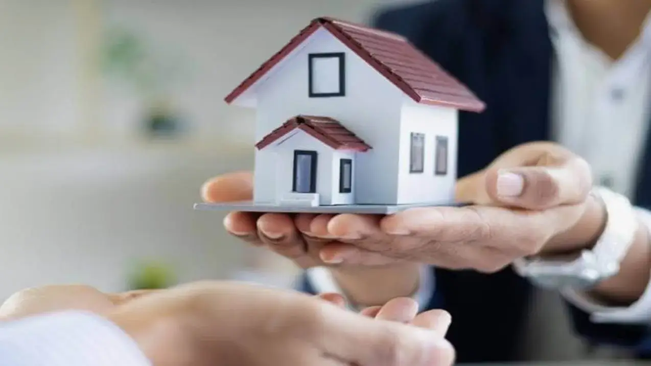 Mão de mulher apresenta uma maquete de casa a outra que se aproxima para fechar negócio. Simbolizando uma Imobiliária de Confiança.