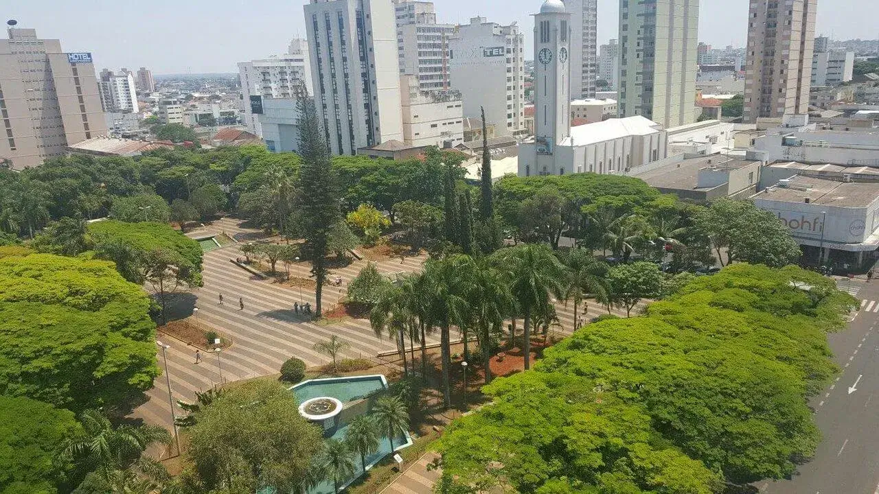 Ângulo e Perspectiva: A imagem é capturada de um ponto de vista elevado, possivelmente de um prédio alto adjacente, proporcionando uma visão abrangente da praça e seu entorno. A perspectiva permite observar a organização da praça, o padrão de caminhos, a densidade da vegetação e a relação entre o espaço verde e o ambiente urbano. Luz e Sombra: A luz solar é predominante, com sombras suaves projetadas pelas árvores e edifícios. A luz destaca a textura das folhas, o padrão dos pavimentos e as fachadas dos edifícios. Atmosfera: A imagem transmite uma sensação de tranquilidade e ordem em meio à cidade. A presença da praça como um espaço verde oferece um contraste com a arquitetura densa e o ritmo da vida urbana. Detalhes da Praça: Vegetação: As árvores variam em tamanho e tipo, desde palmeiras esguias até árvores frondosas com copas densas. A diversidade da vegetação adiciona camadas de verde e textura à praça. Há áreas ajardinadas com arbustos e plantas menores, criando um ambiente convidativo. Caminhos: Os caminhos pavimentados são o destaque da praça, criando um padrão visualmente interessante. O material usado para os caminhos parece ser durável e bem conservado, provavelmente tijolos, pavimentos ou concreto com um acabamento texturizado. Os caminhos são largos o suficiente para permitir que as pessoas caminhem confortavelmente em grupos ou individualmente. Fonte: A fonte é um ponto focal na área da praça. Possui uma estrutura circular com uma bacia de água azul-turquesa. A água jorra do centro da fonte, criando um efeito visual refrescante. A área ao redor da fonte é cuidadosamente ajardinada com palmeiras e plantas menores, criando um espaço agradável para relaxar e apreciar a vista. Iluminação: Há postes de iluminação ao longo dos caminhos, indicando que a praça é usada também à noite. Os postes têm um design simples e funcional, integrando-se ao ambiente sem chamar muita atenção. Detalhes dos Edifícios: Hotel: O prédio do hotel no lado esquerdo da imagem tem uma estrutura imponente, com vários andares e janelas simetricamente dispostas. A placa "HOTEL" no topo é grande e visível de longe. A arquitetura do hotel sugere que é um estabelecimento bem estabelecido. Igreja/Catedral: O edifício branco com a torre do relógio é provavelmente uma igreja ou catedral. A torre do relógio é um marco arquitetônico, com um relógio em cada lado. A arquitetura da igreja é clássica, com linhas limpas e uma fachada imponente. A presença da igreja adiciona um toque de história e cultura ao cenário urbano. Edifícios Residenciais/Comerciais: Os outros edifícios altos ao redor da praça são provavelmente prédios residenciais ou comerciais. A arquitetura desses edifícios é moderna, com fachadas de vidro e concreto. Alguns dos edifícios têm varandas ou terraços, oferecendo vistas da cidade. Loja: No lado direito, o prédio "bohfi" aparenta ser uma loja. Detalhes Adicionais: Tráfego: A rua no canto inferior direito da imagem mostra carros passando, indicando que a praça está localizada em uma área movimentada da cidade. A presença do tráfego adiciona um elemento de dinamismo ao cenário. Céu: O céu claro e azul contribui para a sensação geral de tranquilidade e otimismo. Esta descrição mais detalhada oferece uma visão mais rica e completa da imagem, destacando os elementos-chave e as nuances que a tornam interessante.