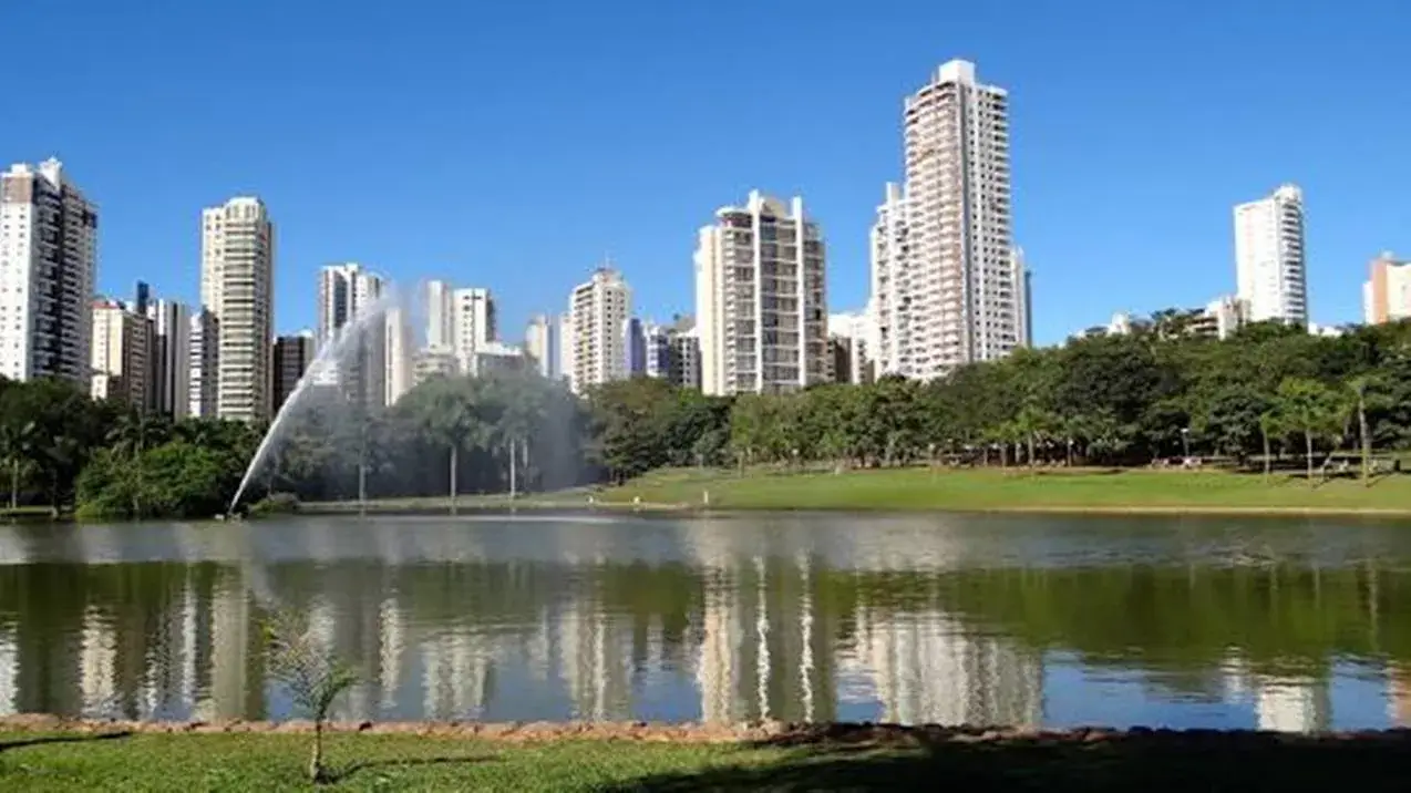 Plano de fundo (parte superior da imagem): Vemos uma linha de prédios altos e modernos, possivelmente residenciais e comerciais, sugerindo um bairro nobre e verticalizado. As construções são majoritariamente brancas ou em tons claros, com design contemporâneo e janelas amplas. O céu está completamente azul, sem nuvens, indicando um dia ensolarado e de clima limpo. Plano intermediário (meio da imagem): Logo à frente dos prédios há uma densa faixa de vegetação, composta por árvores de médio e grande porte. Isso sugere a presença de um parque urbano ou uma área verde bem preservada. Em destaque, há uma fonte d'água lançando um jato alto, que acrescenta movimento e frescor à cena. Algumas pessoas podem ser vistas ao longe, caminhando ou aproveitando o espaço verde, reforçando o aspecto de lazer e convivência. Plano frontal (parte inferior da imagem): Em primeiro plano, há um lago ou espelho d'água, com superfície tranquila, refletindo os prédios, o céu e a vegetação. A margem do lago é gramada e bem cuidada, com um pequeno canteiro ou caminho de terra batida. Há também uma pequena muda de árvore plantada perto da borda do lago, sugerindo cuidados contínuos com o paisagismo. Contexto urbano (provável localização): Essa imagem é muito característica do Setor Bueno, em Goiânia (GO) – um bairro conhecido por sua infraestrutura moderna, grande quantidade de edifícios residenciais e pela presença do Parque Vaca Brava, que parece ser o local retratado na foto. Sensação geral da imagem: A fotografia transmite tranquilidade, organização e qualidade de vida urbana. A combinação de elementos naturais (lago, árvores, céu limpo) com a urbanização vertical (prédios modernos) sugere um local ideal para morar, passear ou praticar atividades ao ar livre.