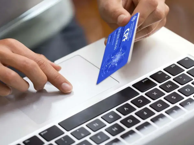 Pessoa em frente ao laptop realiza consulta de imoveis por cpf, com o cartão do documento na mão para verificação.