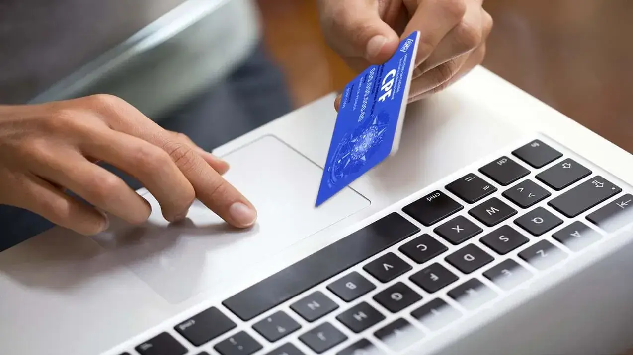 Pessoa em frente ao laptop realiza consulta de imoveis por cpf, com o cartão do documento na mão para verificação.