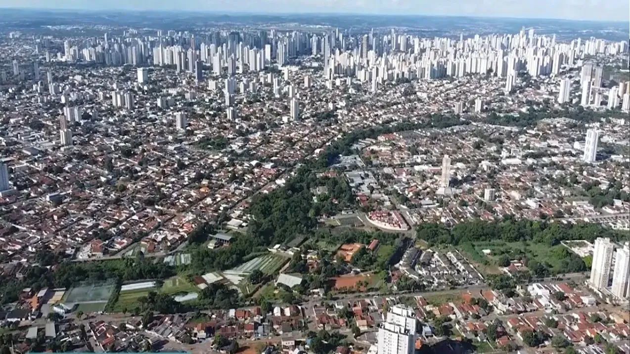 A imagem exibe uma ampla vista aérea panorâmica de uma grande cidade, muito provavelmente Goiânia, sob um céu azul claro e ensolarado. A paisagem urbana se estende até o horizonte, revelando uma metrópole densamente povoada.No fundo e nas laterais da imagem, destaca-se uma concentração significativa de edifícios altos e arranha-céus, predominantemente de cor branca ou clara, formando uma silhueta moderna e verticalizada. Esta área representa o centro ou regiões de maior densidade populacional e desenvolvimento imobiliário da cidade.Em contraste, as áreas no primeiro e médio plano são caracterizadas por uma vasta extensão de bairros residenciais com construções mais baixas. Observa-se uma malha urbana composta por milhares de casas, a maioria com telhados de telha cerâmica (cor de terracota), organizadas em quarteirões e ruas que se entrecruzam. Mesmo nessas áreas mais horizontais, alguns edifícios de apartamentos mais altos se erguem pontualmente, quebrando a uniformidade.Um elemento notável que corta a paisagem diagonalmente é uma extensa faixa de vegetação densa e verde escura. Esta área parece ser um parque linear, um bosque urbano ou uma área de preservação ambiental, possivelmente acompanhando um curso d'água, que serpenteia entre os bairros, oferecendo um respiro natural em meio à estrutura urbana. Próximo a esta faixa verde, no plano mais próximo, são visíveis alguns terrenos cultivados, talvez hortas comunitárias ou viveiros.A fotografia captura a dualidade da cidade: a verticalização intensa em algumas zonas e a predominância de residências unifamiliares em outras, tudo interligado por vias e espaços verdes. A diversidade de moradias e a estrutura da cidade são fatores que impactam diretamente o Custo de Vida em Goiânia, variando significativamente entre os bairros mais verticalizados e os mais tradicionais. A imagem transmite a sensação de uma cidade vibrante, em expansão e com significativa arborização em meio ao concreto.