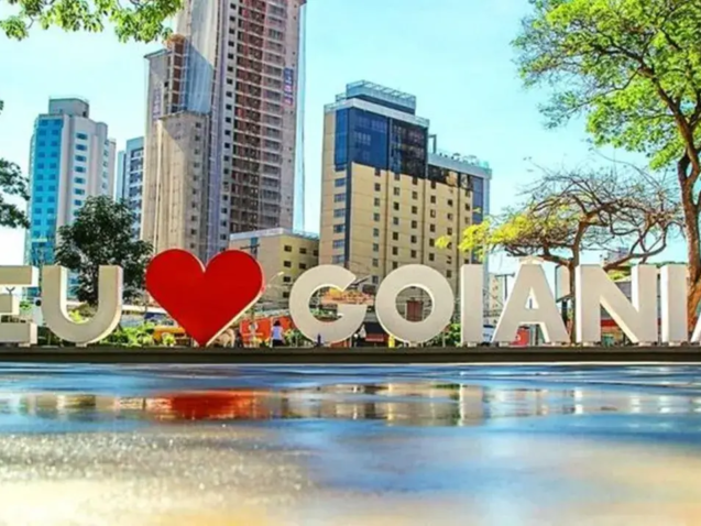 Letreiro "eu amo Goiânia" com coração vermelho, refletido no chão molhado. Ao fundo, prédios e um céu azul em dia ensolarado.