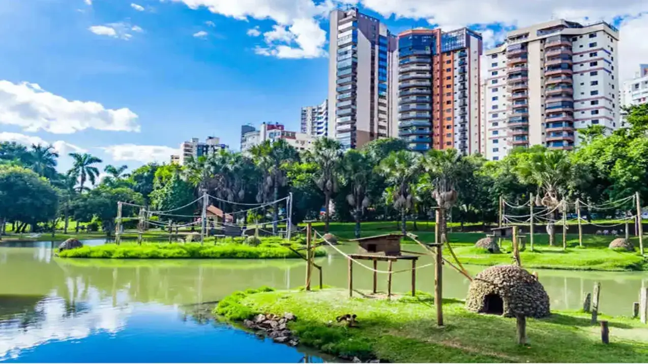 A imagem retrata uma paisagem vibrante em um dia ensolarado, sob um céu azul límpido pontilhado por nuvens brancas e fofas. Ao fundo, ergue-se um conjunto de modernos edifícios altos, arranha-céus predominantemente residenciais com varandas e janelas espelhadas, que compõem parte da linha do horizonte urbana. Suas fachadas variam em cores, incluindo tons de branco, bege, marrom e azul.
Fazendo a transição entre o ambiente urbano e o natural, uma densa faixa de vegetação exuberante, com árvores frondosas de diferentes espécies e diversas palmeiras altas, oferece um verde intenso que contrasta com os prédios.
No plano intermediário e em primeiro plano, um lago de águas calmas e esverdeadas ocupa uma porção significativa da cena, refletindo suavemente o azul do céu, as nuvens e a vegetação circundante. Pequenas ilhas e penínsulas cobertas por grama verde bem aparada se projetam sobre a água, conectadas por estruturas que parecem ser parte de um parque ou zoológico.
Nessas áreas gramadas, notam-se instalações peculiares construídas com materiais naturais como madeira e possivelmente barro ou fibras trançadas. Há pequenas casas de madeira suspensas sobre palafitas, abrigos em formato de cúpula ou colmeia elevados em postes grossos, e passarelas feitas de corda e madeira que interligam algumas dessas estruturas e ilhas, sugerindo um habitat ou área de recreação para animais, possivelmente primatas (pequenos animais podem ser sutilmente vistos em uma das ilhas mais próximas). A grama é vívida e as margens do lago são delineadas por pedras.
A composição geral da imagem cria um contraste interessante entre a natureza (representada pelo parque/zoológico com seu lago e vegetação) e o desenvolvimento urbano (os prédios ao fundo), sugerindo um oásis verde dentro da cidade. A iluminação forte e as cores saturadas indicam um dia de tempo bom e agradável. Esta harmoniosa combinação entre a infraestrutura moderna e espaços verdes bem cuidados contribui para a qualidade de vida, reforçando a percepção de Goiânia Melhor Cidade para se Viver no Brasil.