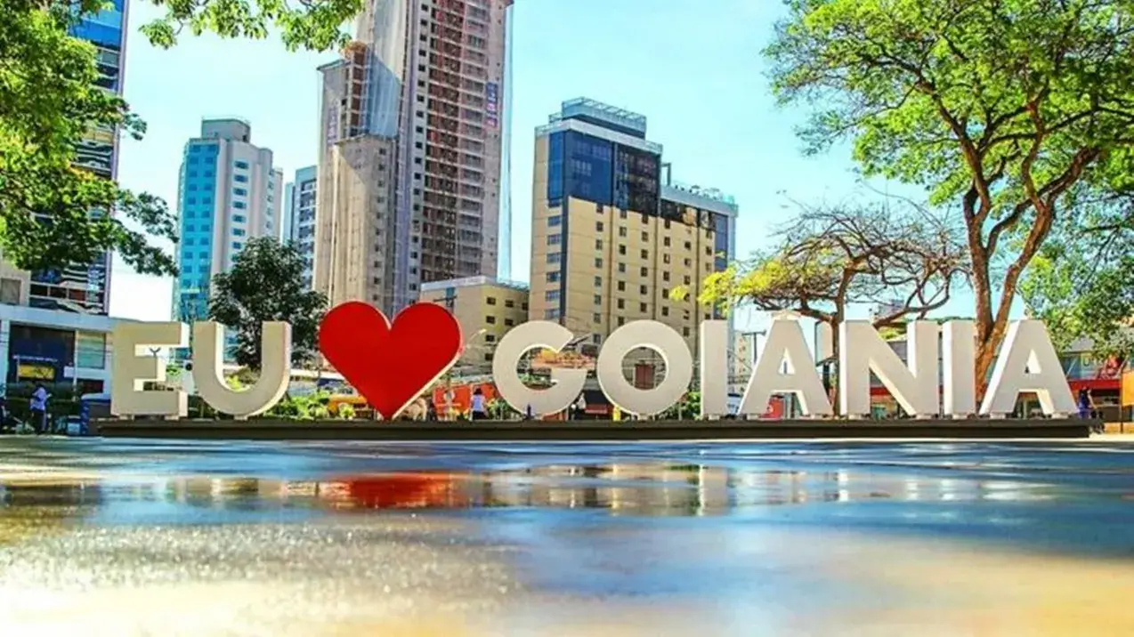 Letreiro "eu amo Goiânia" com coração vermelho, refletido no chão molhado. Ao fundo, prédios e um céu azul em dia ensolarado.