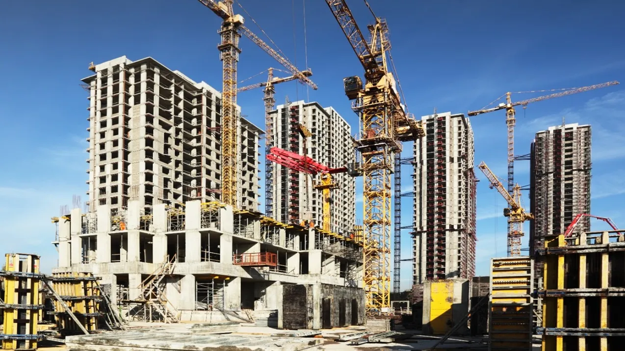 A imagem retrata um grande canteiro de obras, representativo da expansão vista no Mercado Imobiliário em Goiânia, em plena luz do dia, sob um céu azul vibrante e quase sem nuvens. O foco principal são vários edifícios altos (arranha-céus) em diferentes estágios de construção, dominando a paisagem.
Edifícios:
As estruturas são predominantemente de concreto armado, formando esqueletos imponentes, característicos de muitos empreendimentos verticais.
Nenhum revestimento externo, como fachadas ou janelas, foi instalado ainda; as aberturas nas paredes são visíveis, mostrando os vários andares internos.
Os edifícios parecem ser de design moderno e retilíneo, alinhados com as tendências arquitetônicas observadas em novos lançamentos.
Alguns prédios estão mais avançados que outros, mas todos estão claramente na fase estrutural.
Nos andares inferiores de alguns edifícios, e também no nível do solo, podem-se ver cofragens (moldes para concreto), algumas de metal com painéis amarelos e outras de madeira ou metal mais escuro, indicando trabalhos recentes de concretagem ou preparação para futuras etapas.
Vergalhões de aço (barras de reforço) podem ser vistos projetando-se verticalmente de lajes e colunas, aguardando a continuidade da construção para os andares superiores.
Guindastes (Gruas):
Múltiplos guindastes de torre, de cor predominantemente amarela, erguem-se ainda mais altos que os edifícios. Eles estão estrategicamente posicionados entre e ao lado das estruturas.
As lanças (braços) dos guindastes se estendem sobre o canteiro de obras, alguns parecendo inativos no momento da foto, enquanto um guindaste em primeiro plano parece estar em processo de levantar ou posicionar uma grande peça de cofragem de cor vermelha.
A presença de tantos guindastes e a escala dos múltiplos edifícios em construção sugerem a magnitude do projeto, um cenário comum no dinâmico Mercado Imobiliário em Goiânia.
Nível do Solo e Primeiro Plano:
O primeiro plano mostra uma área de trabalho com mais detalhes da construção.
Há lajes de concreto no chão, algumas parecendo ser fundações ou pisos térreos.
Diversos materiais de construção estão espalhados ou empilhados, incluindo mais cofragens, escoras e possivelmente blocos de concreto ou outros elementos pré-fabricados.
A textura geral do solo é áspera, típica de um ambiente de construção.
Iluminação e Atmosfera:
A luz solar é forte, criando sombras definidas, embora não muito longas, sugerindo que pode ser meio da manhã ou meio da tarde.
A imagem transmite uma sensação de atividade industrial intensa, progresso e transformação urbana em grande escala. Apesar da ausência visível de muitos trabalhadores na imagem (talvez alguns pequenos vultos em laranja possam ser vistos em um dos prédios), a cena inteira pulsa com a energia da construção, refletindo o investimento e crescimento característicos do Mercado Imobiliário em Goiânia.
Em resumo, é uma fotografia dinâmica de um complexo de edifícios altos em construção, destacando as estruturas de concreto inacabadas, a presença dominante de guindastes amarelos e a atmosfera de um canteiro de obras ativo sob um céu claro, ilustrando vividamente a atividade do Mercado Imobiliário em Goiânia.
