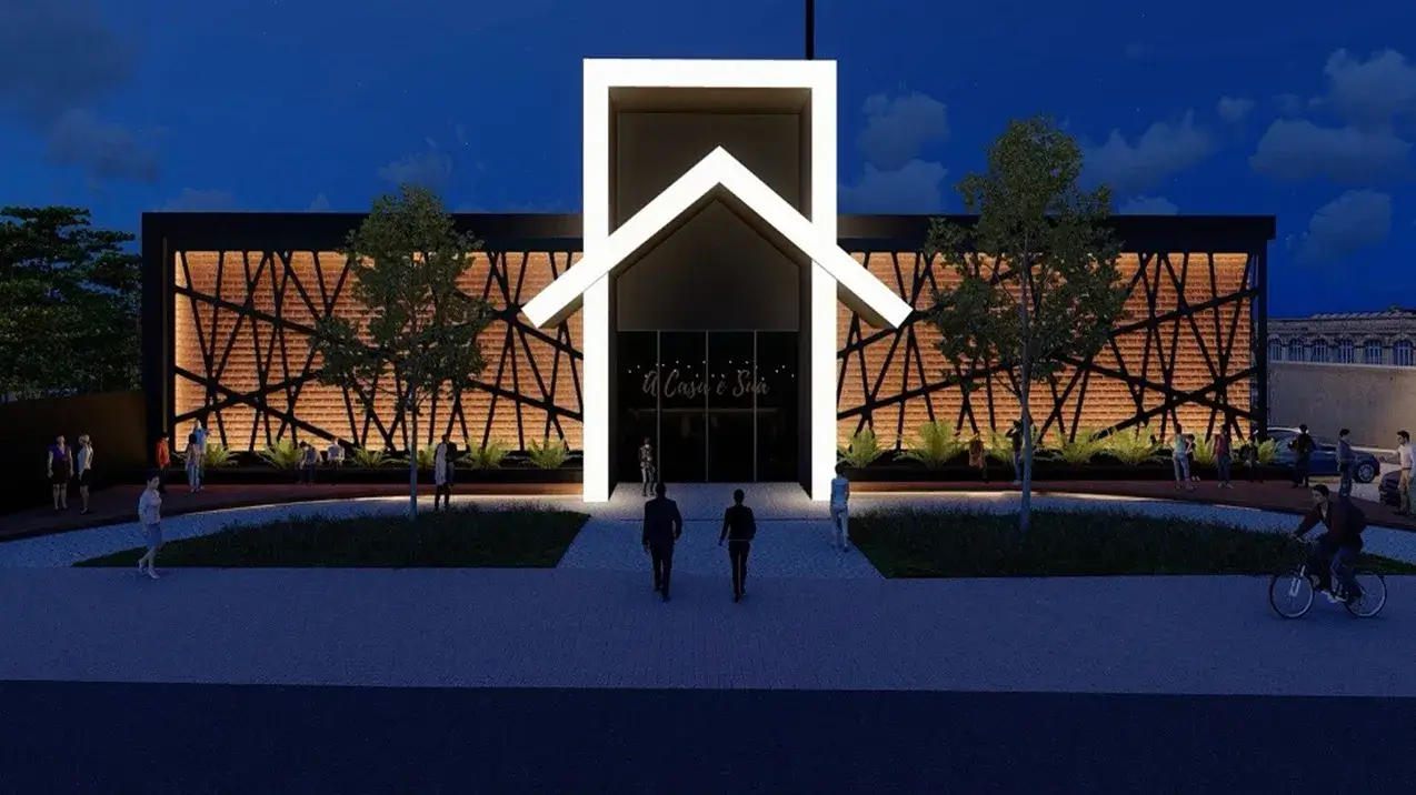 A imagem retrata uma renderização arquitetônica noturna de um edifício moderno com um design marcante. A cena é iluminada artificialmente, destacando as características da construção contra um céu azul escuro de fim de noite com algumas nuvens tênues.
No centro da composição está a fachada do edifício. A entrada principal é proeminente, emoldurada por uma grande estrutura branca e brilhantemente iluminada que tem a forma estilizada do contorno de uma casa – com laterais verticais e um telhado de duas águas no topo. Esta estrutura funciona como um portal luminoso que direciona o olhar para as portas de vidro da entrada. Acima das portas, em letras cursivas e também iluminadas, está escrito "A Casa é Sua".
Flanqueando esta entrada central, as paredes laterais do edifício apresentam uma textura rica, possivelmente de tijolos ou ripas de madeira dispostas horizontalmente, que são retroiluminadas, emitindo um brilho quente e âmbar. Sobre esta superfície texturizada e iluminada, uma estrutura preta com um padrão geométrico complexo, formado por linhas retas que se cruzam em ângulos variados, cria um interessante jogo de luz e sombra, conferindo profundidade e modernidade à fachada. O topo do edifício é reto e escuro, contrastando com a entrada iluminada.
Em frente ao edifício, há uma área pavimentada que serve como acesso. Um caminho central, também pavimentado e de cor clara, leva diretamente à entrada. De cada lado deste caminho, há áreas ajardinadas com grama, algumas árvores de porte médio com folhagem verde e arbustos baixos plantados em canteiros retangulares escuros na base do edifício, iluminados por luzes direcionadas de baixo para cima.
Várias figuras humanas em escala estão distribuídas pela cena, dando uma sensação de vida e atividade ao local. Um casal caminha pelo caminho central em direção à entrada. Outras pessoas estão paradas em pequenos grupos nas laterais, perto das áreas ajardinadas, algumas conversando. Na rua em primeiro plano, que é pavimentada e escura, uma pessoa anda de bicicleta da direita para a esquerda. À direita, parte de um carro estacionado é visível, e mais pessoas estão próximas a ele. Ao fundo, à direita, vislumbra-se parte de uma construção mais antiga, talvez com detalhes em pedra. No lado esquerdo, a cena é delimitada por uma cerca escura e mais vegetação densa.
A iluminação geral é cuidadosamente planejada para realçar a arquitetura, criando um ambiente acolhedor e sofisticado. A combinação da estrutura branca luminosa, das paredes texturizadas com brilho quente e da vegetação iluminada torna o edifício um ponto focal atraente na paisagem noturna, sugerindo que pode ser um centro comunitário, uma igreja moderna, ou um espaço de eventos, representando uma opção interessante sobre o que fazer em Goiânia.