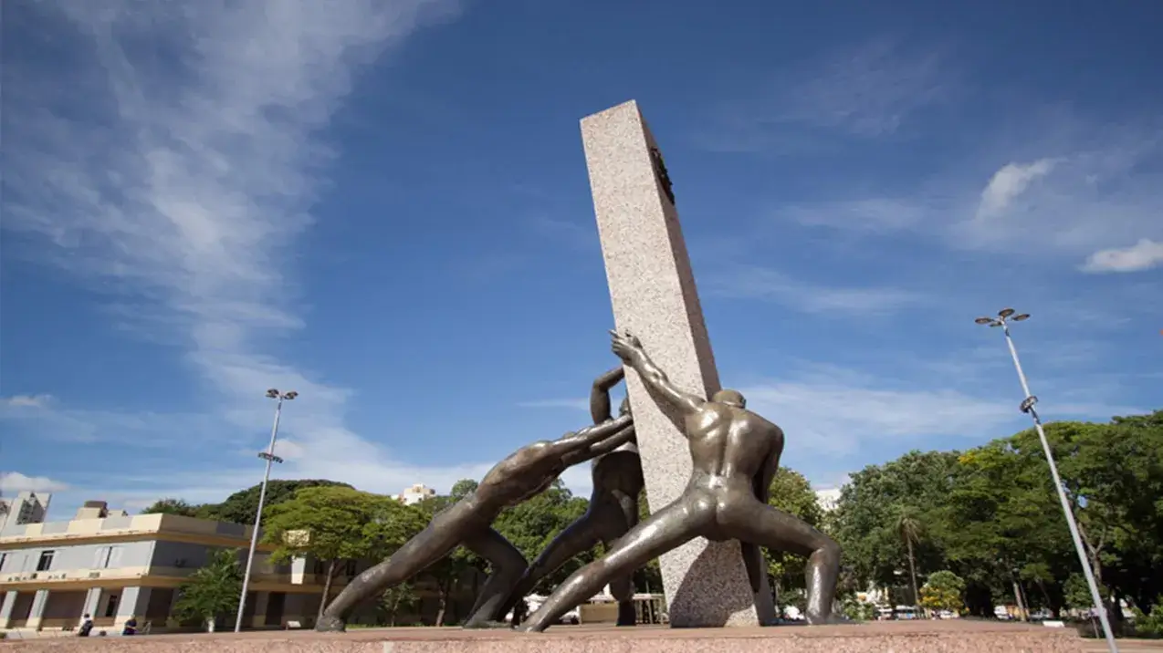 A imagem mostra o imponente Monumento às Três Raças, localizado em Goiânia, sob um céu azul vibrante com nuvens brancas e esparsas, sugerindo um dia ensolarado e agradável. A fotografia é tirada de um ângulo baixo, olhando para cima (contra-plongée), o que acentua a grandiosidade do monumento e a vastidão do céu.
Elemento Central - O Monumento às Três Raças:
Obelisco/Pilar: No centro da composição, ergue-se um grande pilar retangular de pedra clara (provavelmente granito rosado ou similar), com uma textura levemente manchada. Ele está posicionado verticalmente, mas a dinâmica das estátuas ao redor sugere que ele está sendo erguido ou estabilizado por elas.
Estátuas Humanas: Ao redor da base do pilar, três figuras masculinas estilizadas, feitas de bronze ou metal escuro similar com brilho, representam o esforço conjunto. Elas simbolizam as três raças (indígena, negra e branca) que formaram o povo goiano e brasileiro. As figuras estão nuas, com corpos musculosos e definidos, capturadas em poses de extremo esforço físico:
Figura da Direita (mais proeminente): De costas para o espectador, empurra o pilar com as mãos estendidas para cima, pernas afastadas em demonstração de força. A tensão muscular é visível.
Figura da Esquerda: Mais abaixada, empurra o pilar com as costas e ombros, cabeça baixa, corpo totalmente engajado no esforço.
Figura Central (parcialmente visível): Atrás do pilar, também empurra com os braços levantados.
As três figuras juntas transmitem a ideia de união, força e trabalho na construção ou fundação de algo.
Base: O monumento assenta sobre uma base elevada de cor clara (concreto ou pedra), separando-o do nível do chão da praça.
Plano de Fundo e Arredores:
Céu: Ocupa grande parte da imagem, azul intenso com nuvens brancas, finas e alongadas.
Vegetação: Árvores com copas verdes e densas à direita e parcialmente à esquerda indicam um ambiente de praça ou parque (Praça Cívica).
Edificações: Ao fundo à esquerda, parte de um edifício claro de vários andares é visível. Outras estruturas urbanas se misturam à paisagem.
Postes de Iluminação: Dois postes de luz são visíveis, um à esquerda e outro mais alto e proeminente à direita.
Ambiente: O cenário é um espaço público aberto. Pequenas figuras humanas sentadas na base ao longe dão escala ao Monumento às Três Raças.
Iluminação e Atmosfera:
A luz solar incide da direita e de cima, criando sombras marcadas e realçando os volumes e texturas do pilar e das estátuas de bronze. A atmosfera geral, reforçada pelo nome Monumento às Três Raças, é de força, esforço coletivo, fundação, união e celebração da miscigenação e da construção de uma identidade.