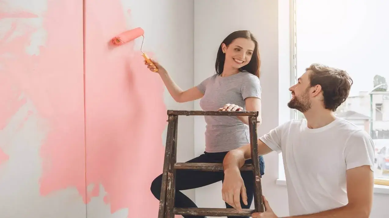 Casal em reforma. A mulher, numa escada, pinta a parede de rosa, enquanto o homem, no chão, a observa sorrindo com admiração e cumplicidade.