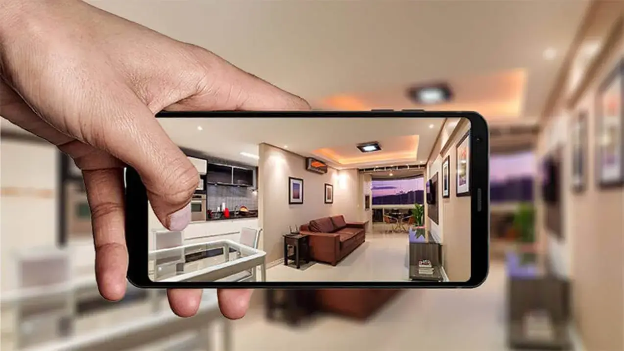 Mão segura um celular exibindo a foto de um apartamento moderno, com sala de estar, piso de madeira e um sofá de couro.