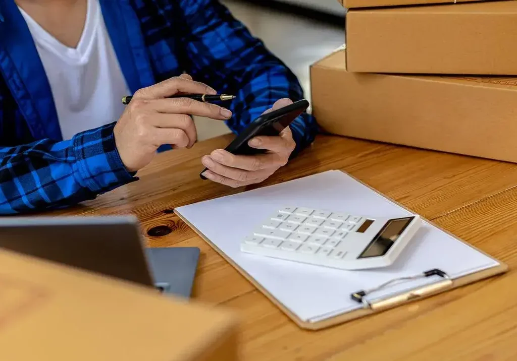 Homem de camisa xadrez calcula em seu celular os custos para o Aluguel de Box em Goiânia, com caixas ao lado em uma mesa.