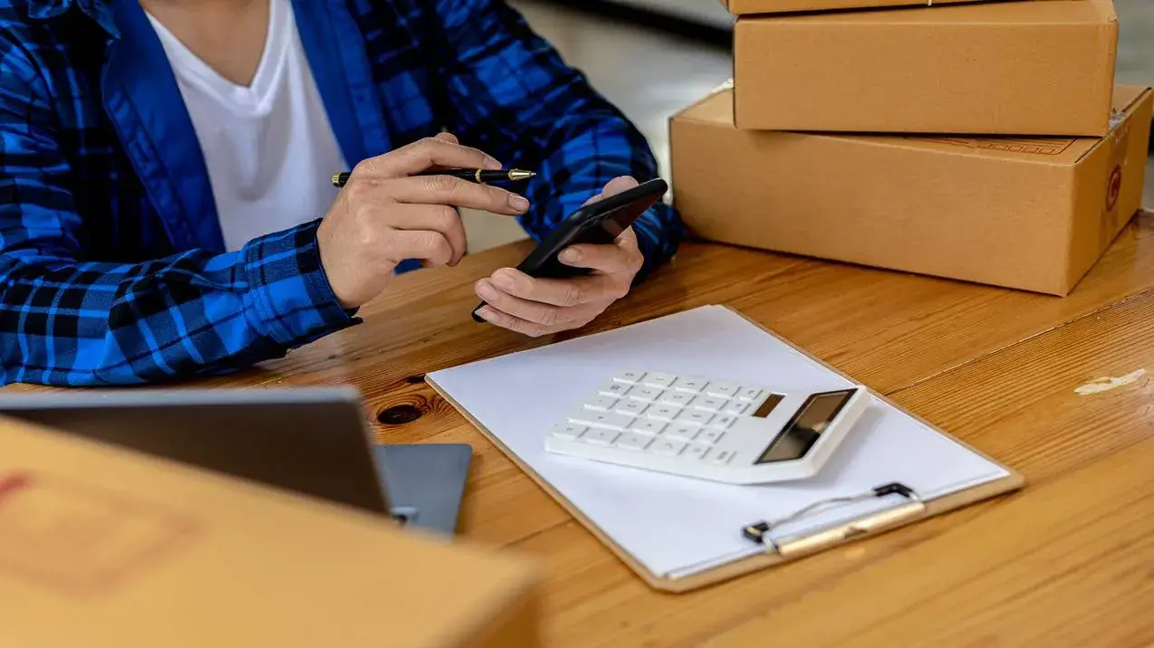 Homem de camisa xadrez calcula em seu celular os custos para o Aluguel de Box em Goiânia, com caixas ao lado em uma mesa.
