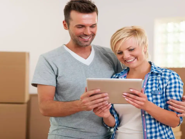 Casal feliz planejando o custo do guarda móveis em Goiânia usando um tablet, com caixas de mudança ao fundo.