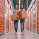 Jovem casal sorridente carrega caixas em um corredor de self storage com boxes laranja. Solução de guarda móveis.